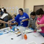 Participants at the table are engaged in the VentureLab Idea to Pitch program, sorting bags, water bottles, and papers as part of the implementation process.