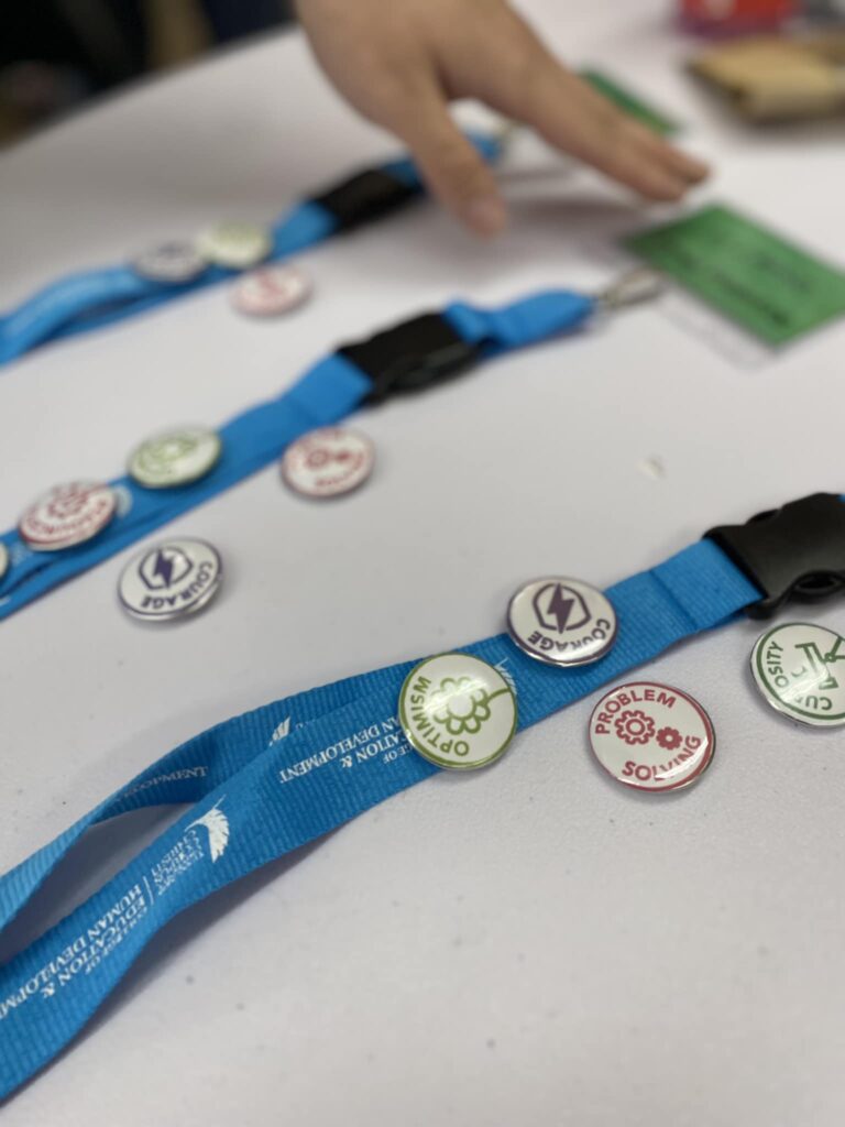 Blue lanyards with various buttons labeled "Problem Solving," "Forward," and more lay on a table, part of the VentureLab Idea to Pitch program implementation. A hand reaches toward a green object.