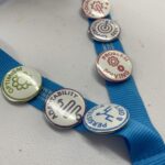 Five buttons with words: "Optimism," "Adaptability," "Resourcefulness," "Problem Solving," and "Persistence & Grit" adorn a blue ribbon, symbolizing the core values driving the VentureLab Idea to Pitch program implementation.