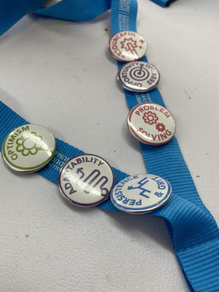 Five buttons with words: "Optimism," "Adaptability," "Resourcefulness," "Problem Solving," and "Persistence & Grit" adorn a blue ribbon, symbolizing the core values driving the VentureLab Idea to Pitch program implementation.