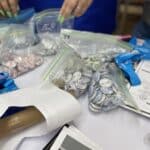 Two people meticulously sort button pins and lanyards into plastic bags as part of the VentureLab Idea to Pitch program implementation, their table cluttered with paper and pens, each item representing a small step toward bringing innovative visions to life.