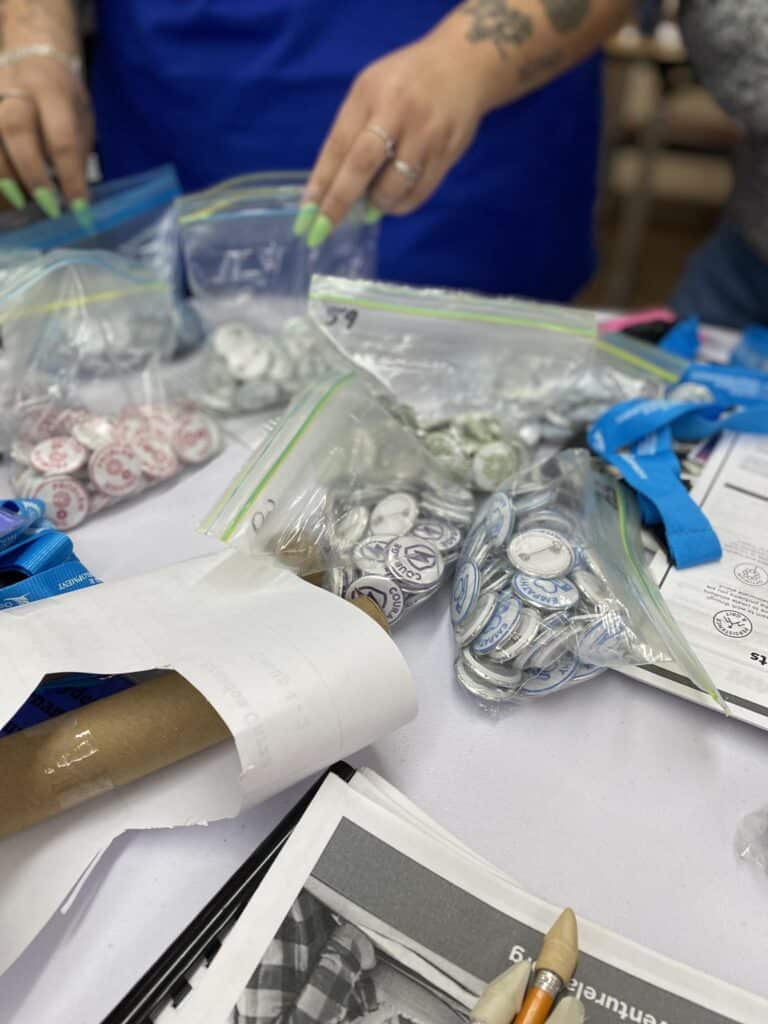 Two people meticulously sort button pins and lanyards into plastic bags as part of the VentureLab Idea to Pitch program implementation, their table cluttered with paper and pens, each item representing a small step toward bringing innovative visions to life.