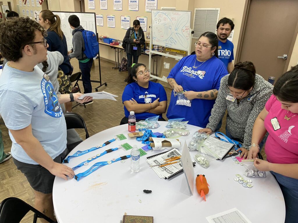 People at a table discussing the VentureLab Idea to Pitch program implementation; some are wearing blue "TRIO" shirts. Various items, including buttons and lanyards, are spread out on the table.