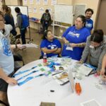 People at a table discussing the VentureLab Idea to Pitch program implementation; some are wearing blue "TRIO" shirts. Various items, including buttons and lanyards, are spread out on the table.