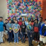 A group of people stands in front of a colorful flower wall, some proudly displaying lanyards from the VentureLab Idea to Pitch program.