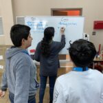 Three students stand in front of a whiteboard preparing for the VentureLab Idea to Pitch program. One writes "Being eaten" under a "Cons" list, while two others watch. The board also boasts a "Pros" list with several points ready for implementation.