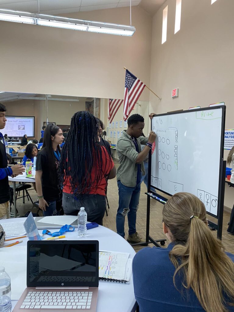 In the Garcia Center classroom, a group gathers around a whiteboard where one participant writes as others observe. An American flag stands in the background, symbolizing unity and learning during the Spring Break Camp Youth Entrepreneurship program.