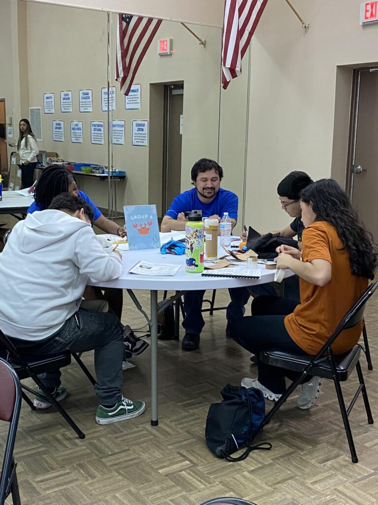In a room adorned with American flags and posters, people gather around a table with snacks and a sign labeled "Group 6" during the student pitch event at the Garcia Center.