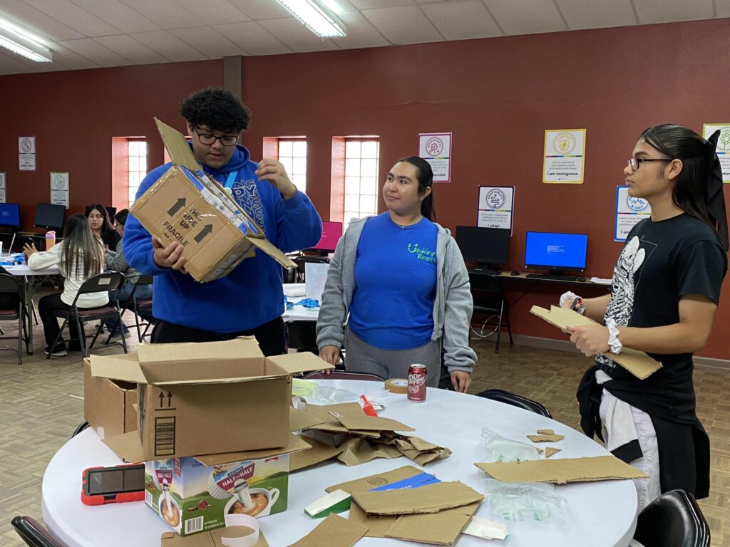 Three people assemble cardboard boxes at a round table in a room buzzing with computers, preparing for the upcoming student pitch event at the Garcia Center.