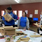 Three people assemble cardboard boxes at a round table in a room buzzing with computers, preparing for the upcoming student pitch event at the Garcia Center.