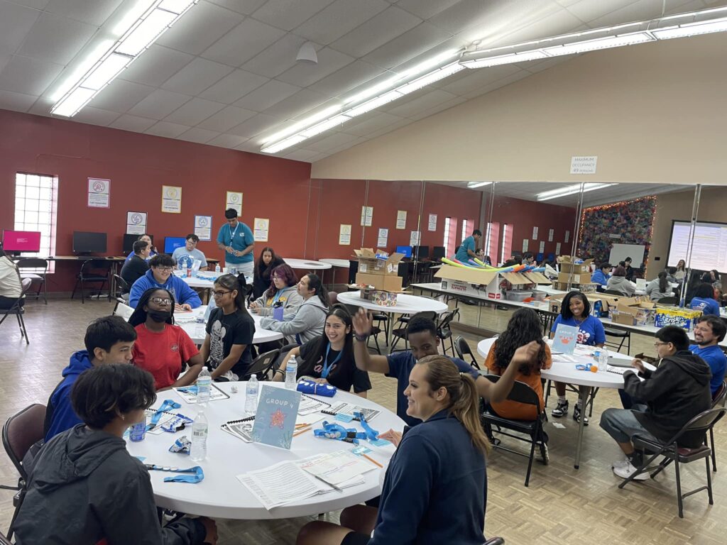 During a vibrant spring break camp, a group of people sits at tables in a lively room, engaged in activities and discussions, with various materials and signs on the tables.