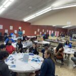 During a vibrant spring break camp, a group of people sits at tables in a lively room, engaged in activities and discussions, with various materials and signs on the tables.