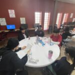 A group of people sitting around a table, deeply engaged in discussion for an upcoming idea to pitch. Computers hum softly in the background, setting the stage for creativity and collaboration.