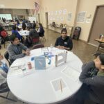 At the Garcia Center's student pitch event, a group of people sit around a round table, engaging in lively discussion. With papers and drinks before them and the American flag proudly displayed in the background, they embrace near peer mentoring to inspire and innovate.