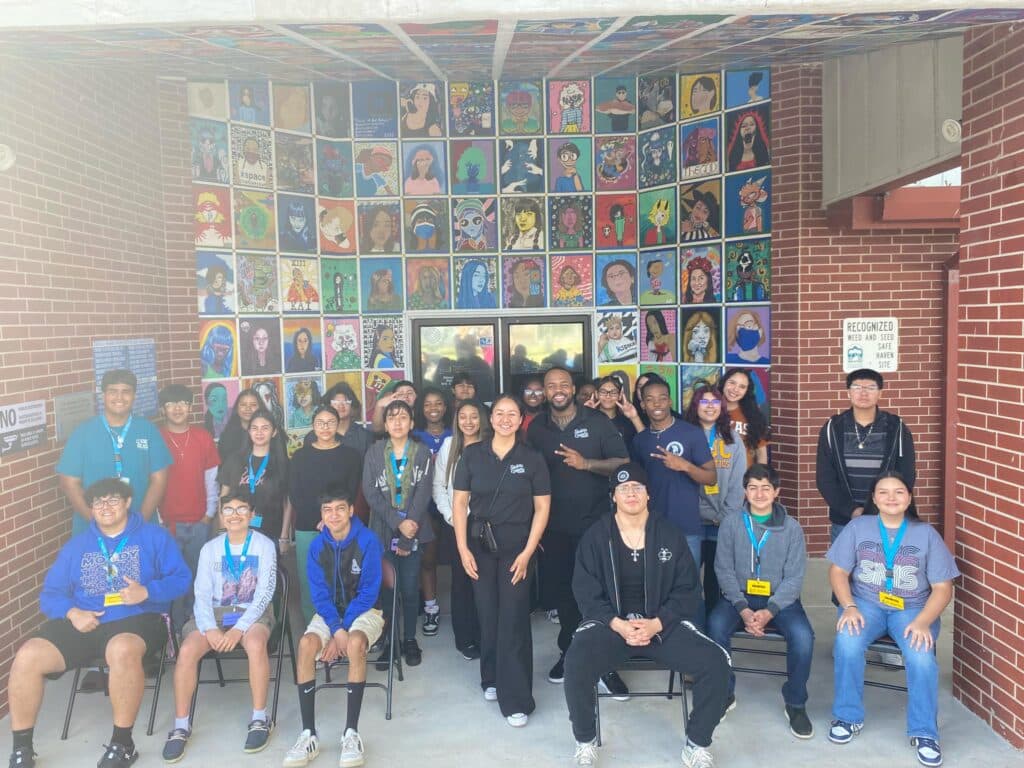 A group of people, including students and staff, posing in front of a wall covered with colorful artwork, showcases the enthusiasm from the Garcia Center Spring Break Camp Youth Entrepreneurship program.