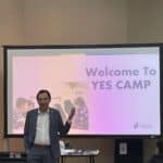 A person in a suit is speaking in front of a screen displaying "Welcome to YES CAMP," featuring the Garcia Center logo. This student pitch event transforms youthful creativity from idea to pitch, set in a room equipped with a projector and tables, perfect for aspiring entrepreneurs.