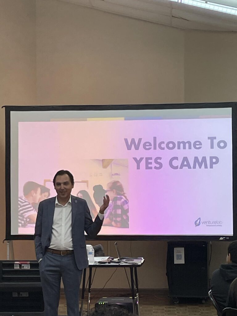 A person in a suit is speaking in front of a screen displaying "Welcome to YES CAMP," featuring the Garcia Center logo. This student pitch event transforms youthful creativity from idea to pitch, set in a room equipped with a projector and tables, perfect for aspiring entrepreneurs.