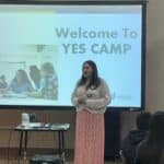 A woman stands before a screen displaying "Welcome to YES Camp," addressing the audience at the Garcia Center Spring Break Camp. Nearby, a projector and table are ready for her to turn ideas into pitches.