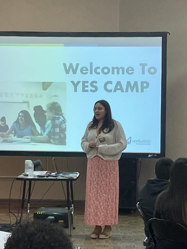 A woman stands before a screen displaying "Welcome to YES Camp," addressing the audience at the Garcia Center Spring Break Camp. Nearby, a projector and table are ready for her to turn ideas into pitches.