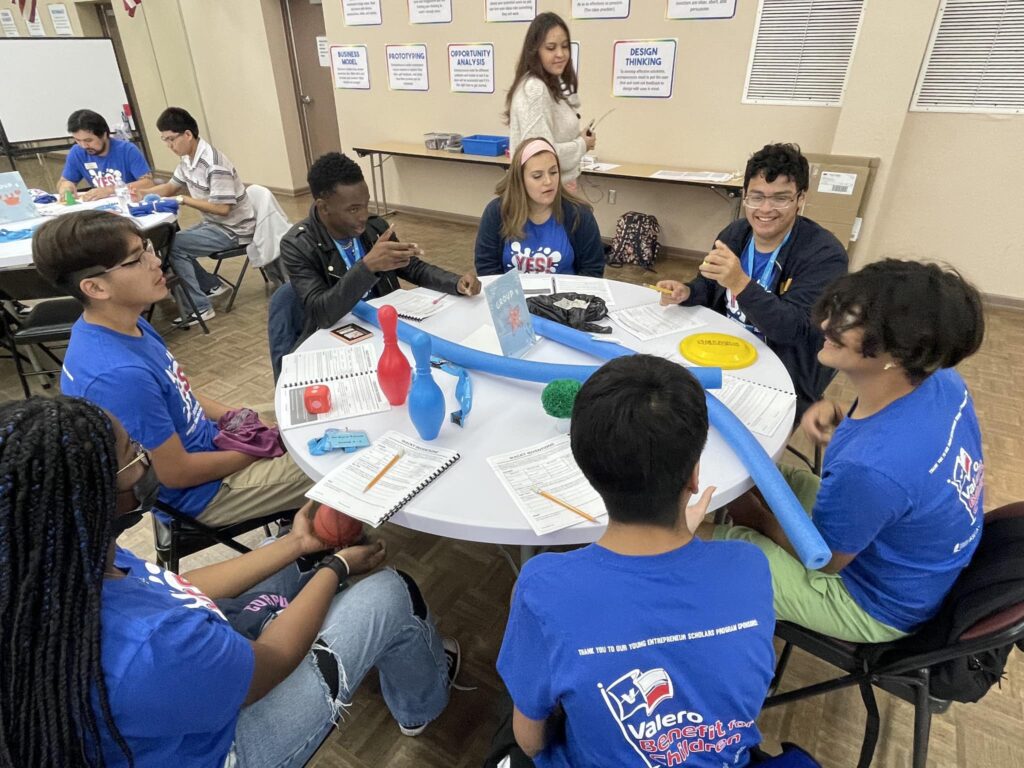 At the Garcia Center, a group of students in blue shirts gathers around a table with notebooks and toy bowling pins, enthusiastically preparing their ideas to pitch for the upcoming student pitch event.