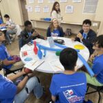 At the Garcia Center, a group of students in blue shirts gathers around a table with notebooks and toy bowling pins, enthusiastically preparing their ideas to pitch for the upcoming student pitch event.