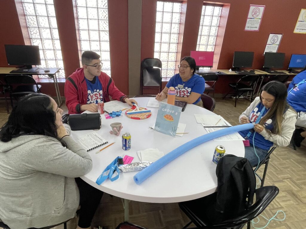 Four people sit around a table, surrounded by notebooks, a pool noodle, snacks, and drinks. They're deep in discussion at the Garcia Center, with computers in the background—perhaps planning for an upcoming spring break camp or coordinating near peer mentoring sessions.
