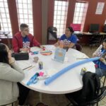 Four people sit around a table, surrounded by notebooks, a pool noodle, snacks, and drinks. They're deep in discussion at the Garcia Center, with computers in the background—perhaps planning for an upcoming spring break camp or coordinating near peer mentoring sessions.