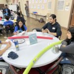 Students at the spring break camp gather around a round table with activity materials, including colored tubes and paper. A sign reads "Group 1," setting the stage for their Idea to Pitch session.