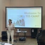 A person enthusiastically presents in front of a screen that reads "Welcome to YES Camp," setting the stage for a dynamic student pitch event. The engaged audience, seated in the room, is ready to explore innovative ideas and experience near peer mentoring.