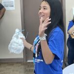 Woman in a blue shirt, smiling and holding a white decorative object, waves her hand in a room with a brown door. She exudes enthusiasm as if she's just returned from Garcia Center Spring Break Camp Youth Entrepreneurship, ready to turn her idea into a pitch.