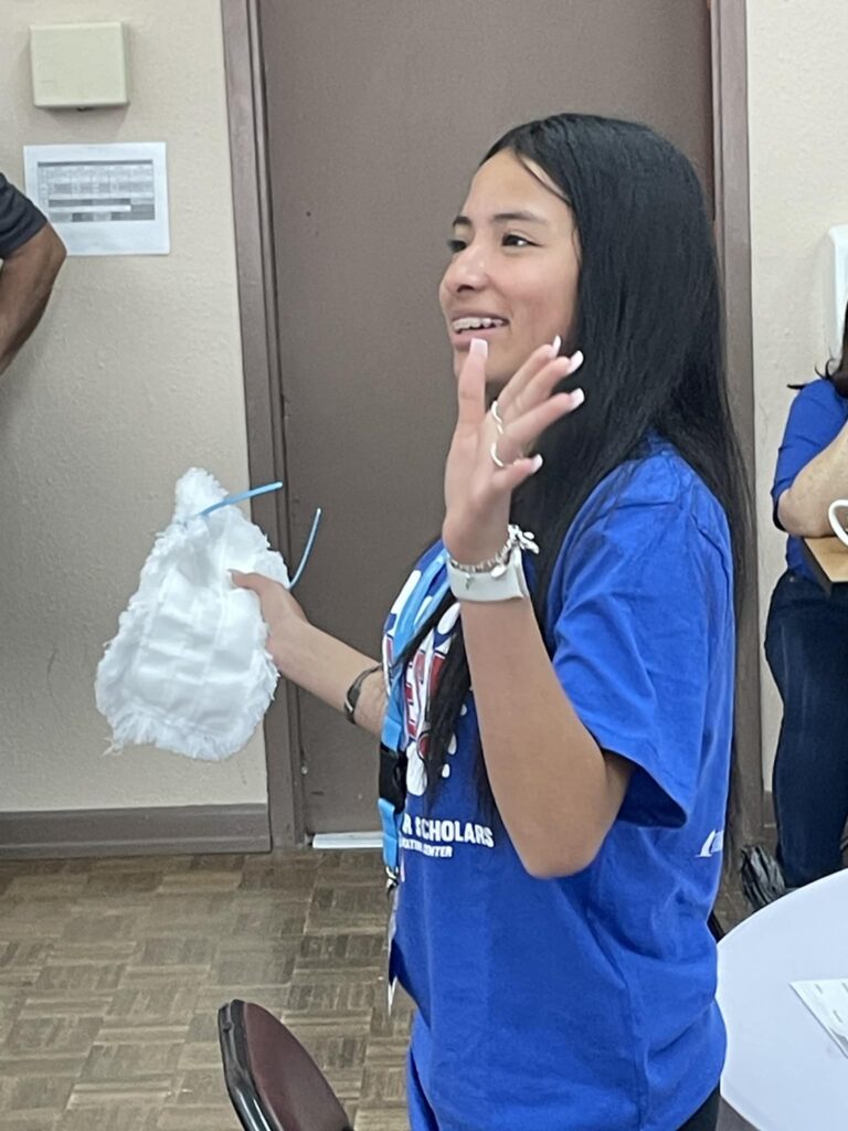 Woman in a blue shirt, smiling and holding a white decorative object, waves her hand in a room with a brown door. She exudes enthusiasm as if she's just returned from Garcia Center Spring Break Camp Youth Entrepreneurship, ready to turn her idea into a pitch.