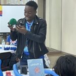 At the Garcia Center, a person in a leather jacket holds a green object, smiling brightly at a group during the vibrant spring break camp. The room buzzes with energy, decorated with flags and colorful items on tables, fostering an inspiring near peer mentoring environment.