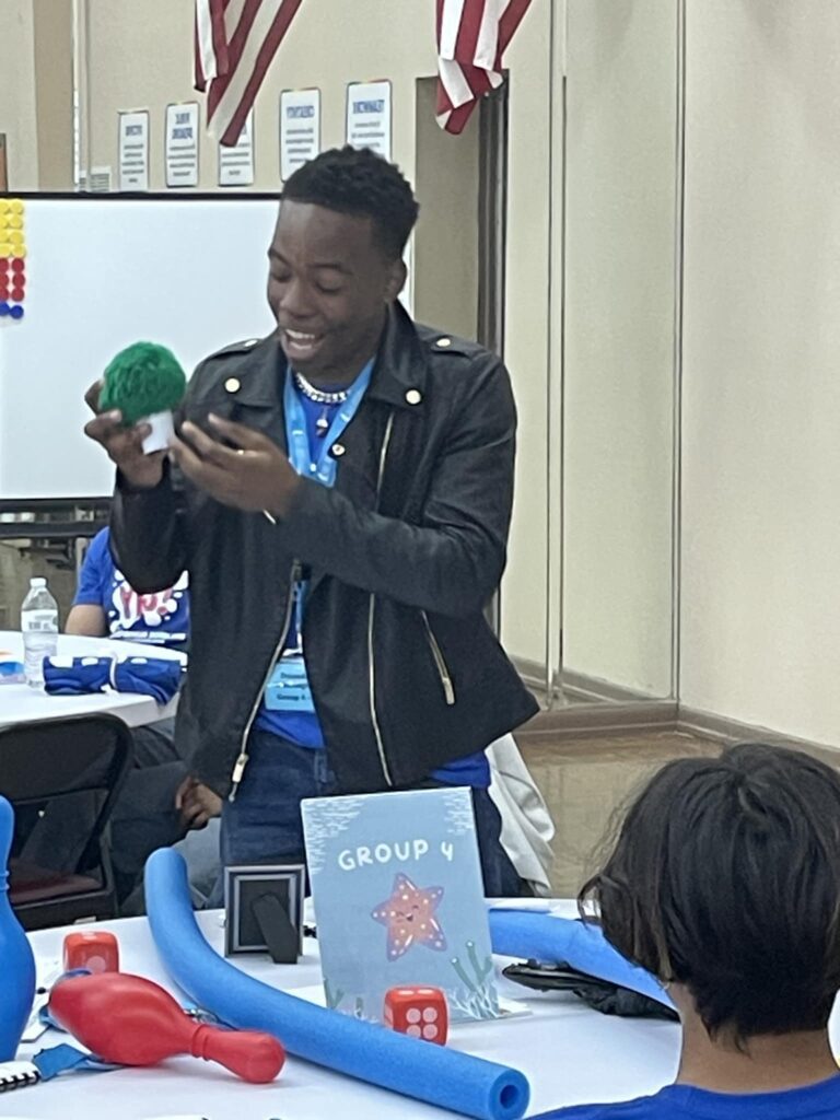 At the Garcia Center, a person in a leather jacket holds a green object, smiling brightly at a group during the vibrant spring break camp. The room buzzes with energy, decorated with flags and colorful items on tables, fostering an inspiring near peer mentoring environment.