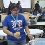 A person in a blue shirt stands at a table brimming with craft supplies in the lively classroom of Garcia Center's Spring Break Camp. In the background, a projection screen flickers, while excited participants prepare for an upcoming student pitch event.