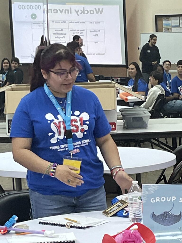 A person in a blue shirt stands at a table brimming with craft supplies in the lively classroom of Garcia Center's Spring Break Camp. In the background, a projection screen flickers, while excited participants prepare for an upcoming student pitch event.
