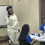 A person in a white outfit stands smiling and holding a phone, ready to pitch an idea, while others sit at tables with papers and drinks. An American flag is visible in the background, capturing the essence of youth entrepreneurship at Garcia Center Spring Break Camp.