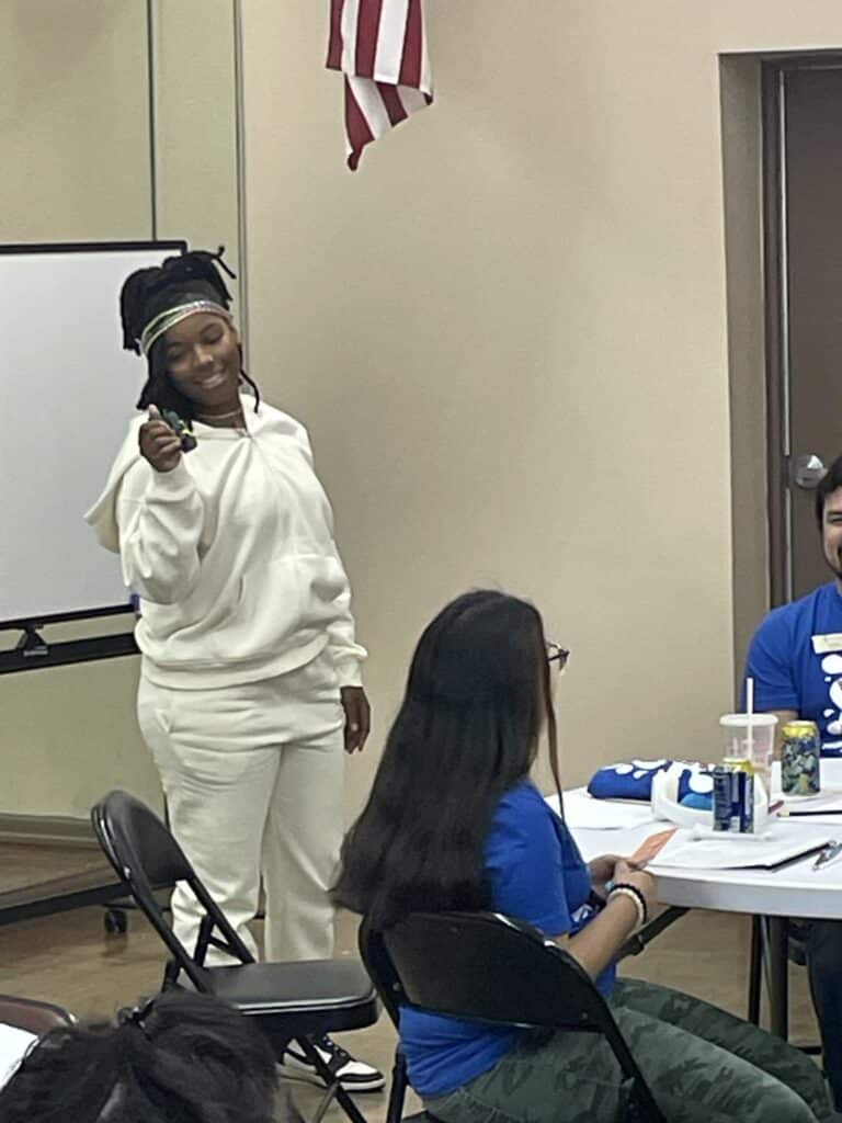 A person in a white outfit stands smiling and holding a phone, ready to pitch an idea, while others sit at tables with papers and drinks. An American flag is visible in the background, capturing the essence of youth entrepreneurship at Garcia Center Spring Break Camp.