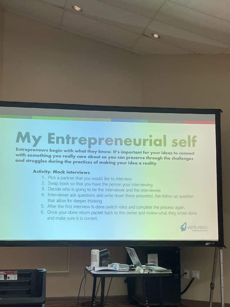 The projector screen displays a slide titled "My Entrepreneurial Self" with steps for a mock interview activity, part of the Garcia Center Spring Break Camp Youth Entrepreneurship program. In front of the screen, a table filled with office supplies awaits eager participants ready to turn their ideas to pitches.