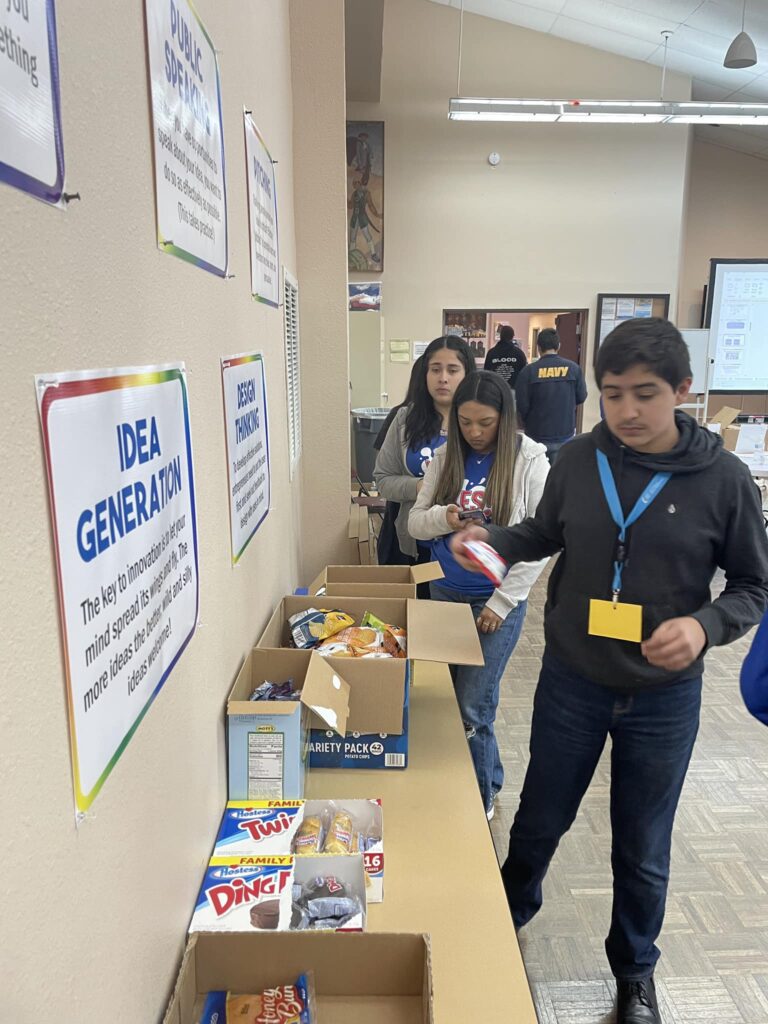 People sorting snacks into boxes on a table at the Garcia Center, surrounded by posters about idea generation, create an inspiring scene. This activity is part of the Garcia Center Spring Break Camp Youth Entrepreneurship program, fostering learning through near peer mentoring.