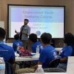 During the spring break camp at Garcia Center, a person presents "Empowered Youth Anatomy Course: What is Stress?" to a seated audience wearing blue shirts in a classroom.