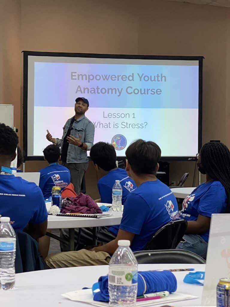 During the spring break camp at Garcia Center, a person presents "Empowered Youth Anatomy Course: What is Stress?" to a seated audience wearing blue shirts in a classroom.