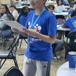 A person in a blue shirt stands confidently, holding papers and speaking passionately at the Garcia Center's Idea to Pitch event, engaging a room filled with seated individuals and tables.