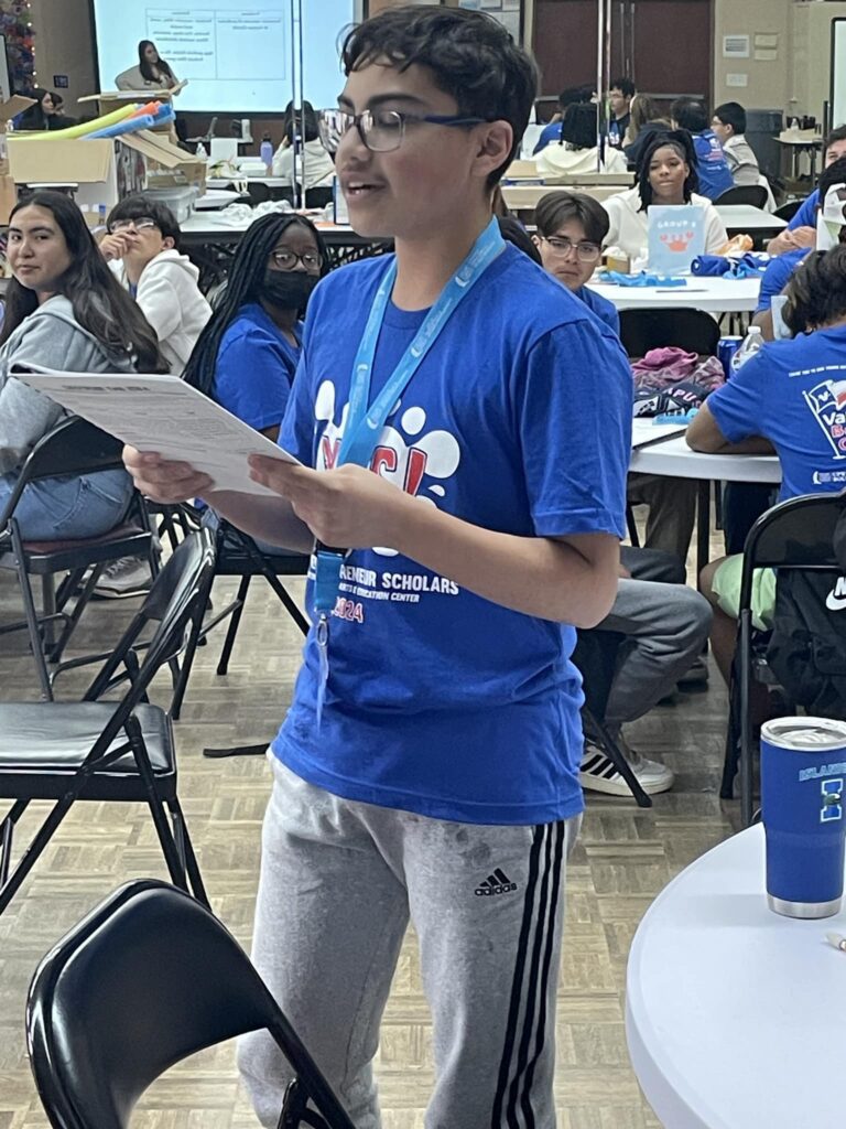 A person in a blue shirt stands confidently, holding papers and speaking passionately at the Garcia Center's Idea to Pitch event, engaging a room filled with seated individuals and tables.