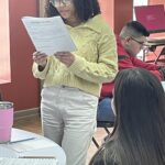 A person in a yellow sweater stands reading a document at the Garcia Center, while others sit at tables with laptops and notes, preparing their Idea to Pitch for the upcoming spring break camp.