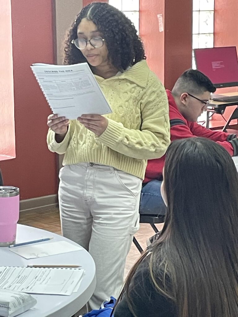 A person in a yellow sweater stands reading a document at the Garcia Center, while others sit at tables with laptops and notes, preparing their Idea to Pitch for the upcoming spring break camp.