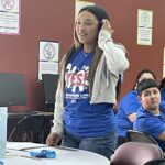 A person stands in a classroom, passionately speaking about the "Idea to Pitch" program while wearing a blue "YES!" shirt and gray hoodie. Posters of the Garcia Center Spring Break Camp Youth Entrepreneurship line the walls, emphasizing near-peer mentoring opportunities.