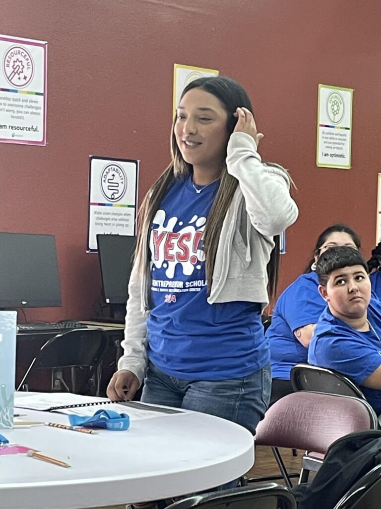 A person stands in a classroom, passionately speaking about the "Idea to Pitch" program while wearing a blue "YES!" shirt and gray hoodie. Posters of the Garcia Center Spring Break Camp Youth Entrepreneurship line the walls, emphasizing near-peer mentoring opportunities.