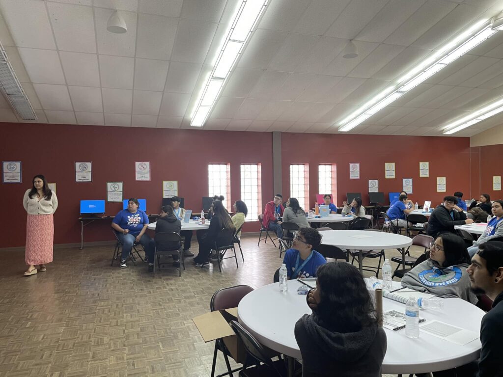 A group of people sit at round tables in a large room with a red wall. A woman stands in front, leading a student pitch event near the presentation screen.