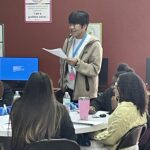 At the Garcia Center, a person stands and reads a paper at a table surrounded by several seated people, engaging in near peer mentoring during the Spring Break Camp Youth Entrepreneurship program.