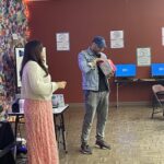 A man in a denim jacket and cap stands next to a woman in a sweater and long skirt, looking into a gift bag in a room with colorful decorations and computers, reminiscent of a student pitch event.